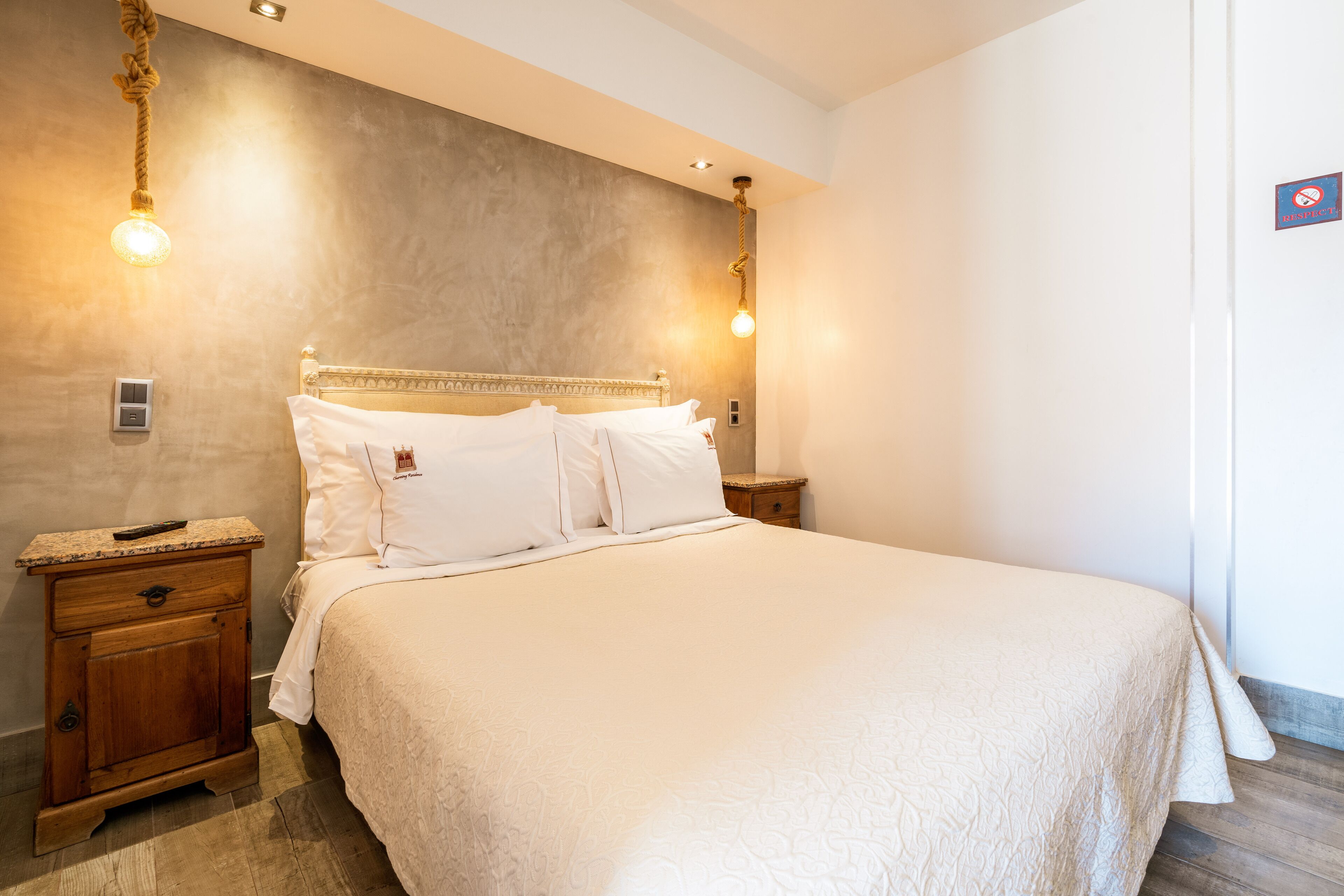 A neatly made bed with white linens, a wooden nightstand with a remote control, and a wall-mounted light fixture.