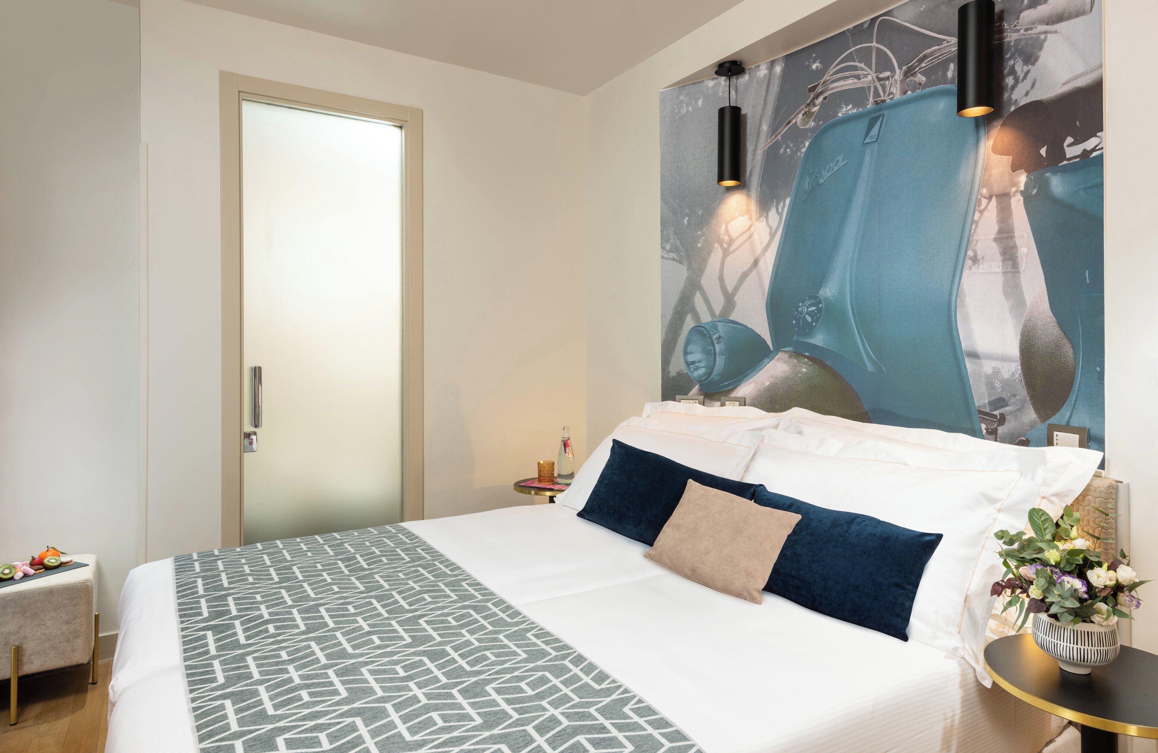 Quarto de hotel moderno com uma cama grande, uma mesinha, um criado-mudo, luminária fixada na parede e uma obra de arte emoldurada sobre a cama.