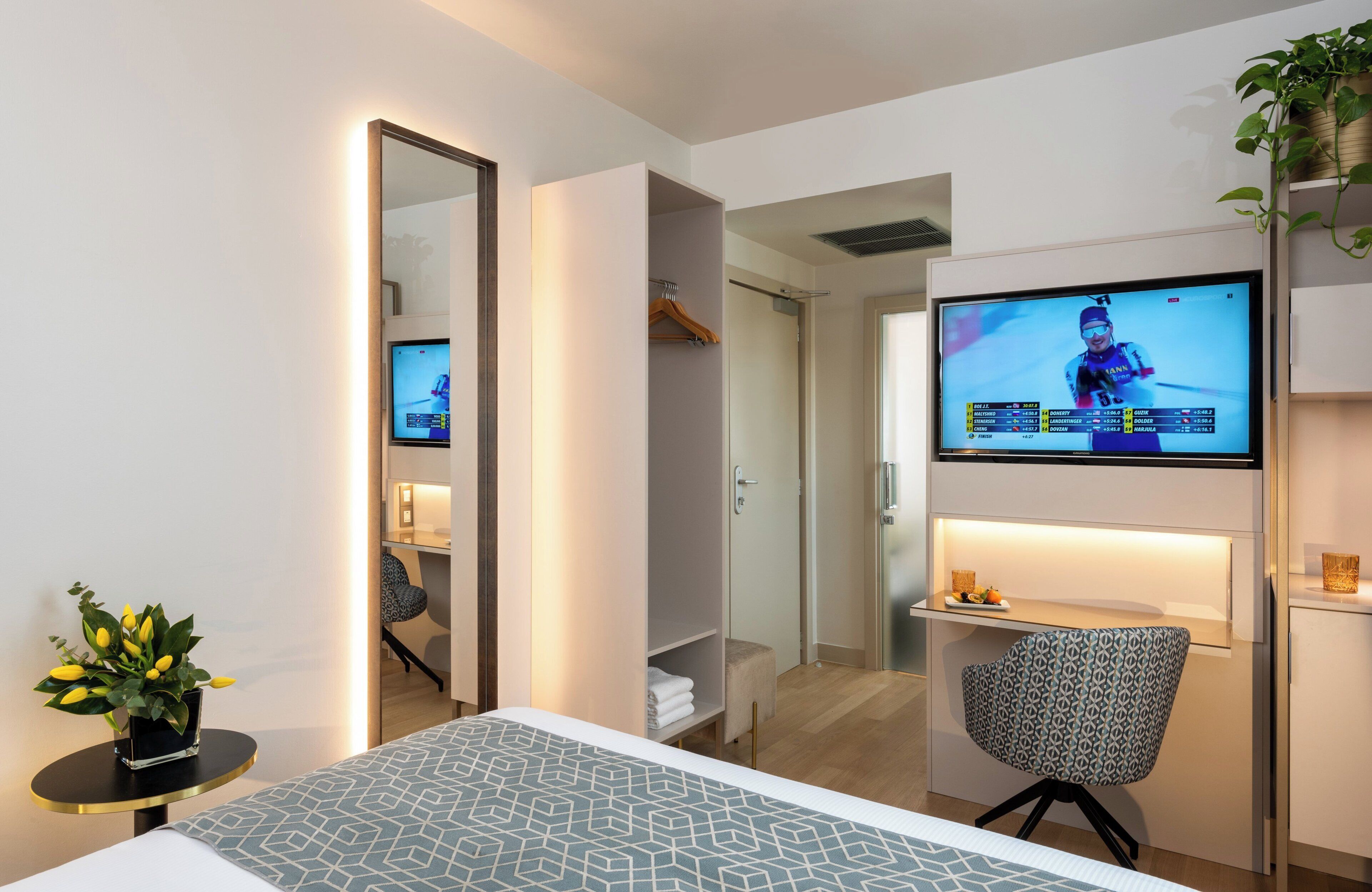 Habitación de hotel con cama, televisor montado en la pared, escritorio con silla, espejo y planta en maceta.