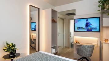 Habitación de hotel con cama, televisor montado en la pared, escritorio con silla, espejo y planta en maceta.