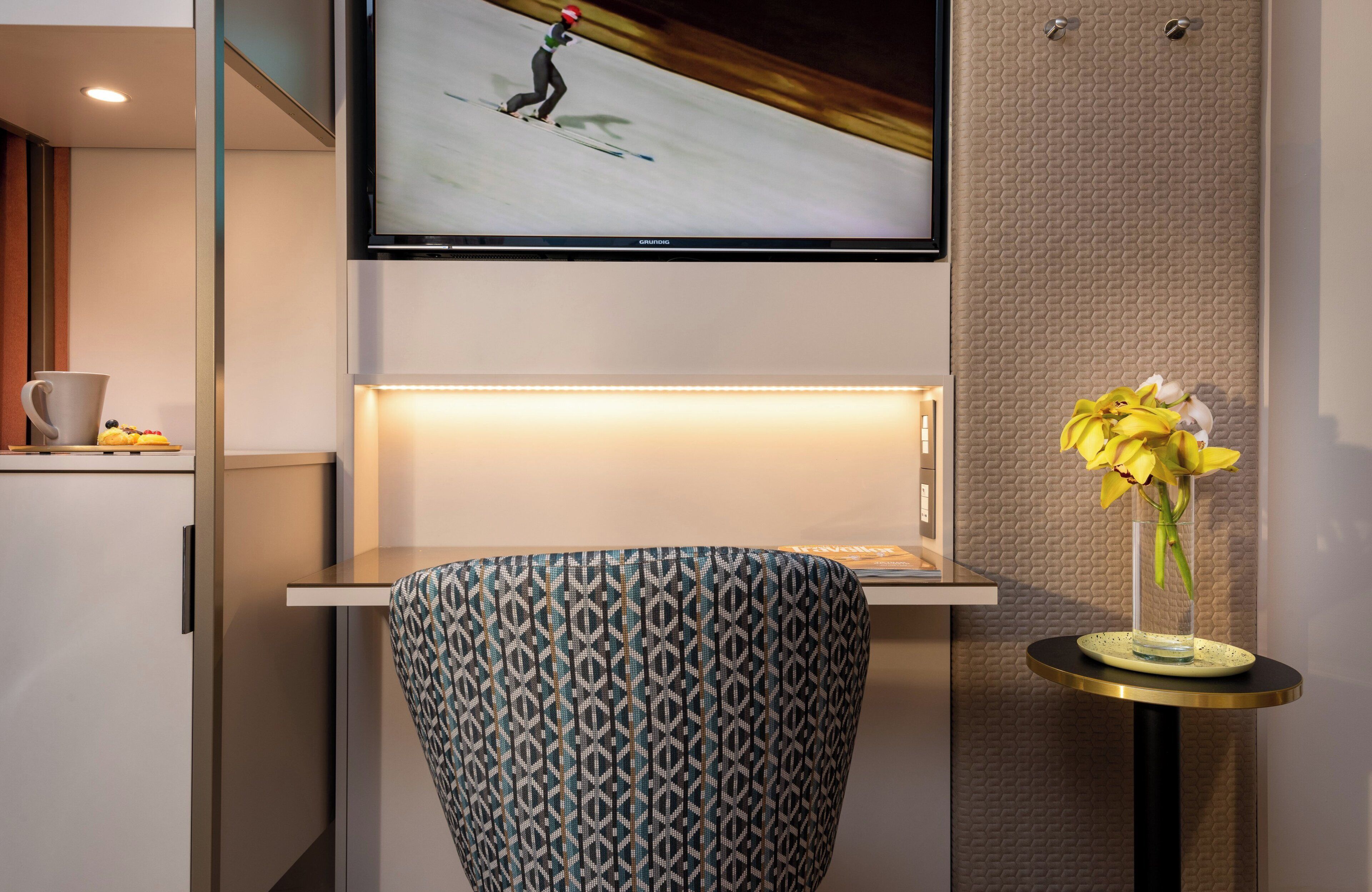 Een moderne hotelkamer met een flatscreen televisie aan de muur, een gestreept fauteuil, een klein tafeltje met een bloemenvaas, en een kopje op een bijzettafeltje.