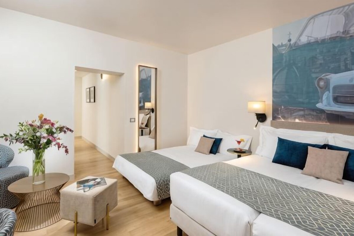 A hotel room with two beds, a sitting area with a round table, a mirror, and a painting on the wall.