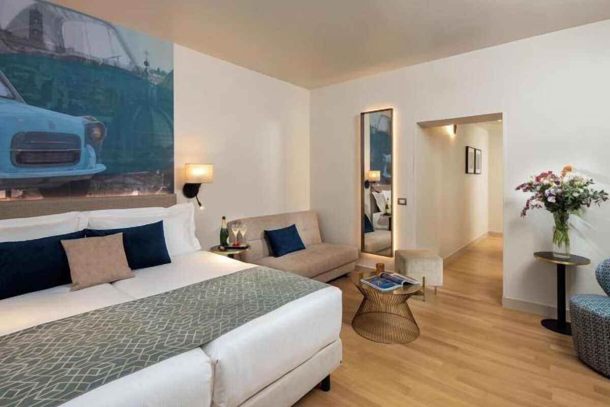A hotel room with a bed, a sofa, a small table, a mirror, and a vase with flowers.