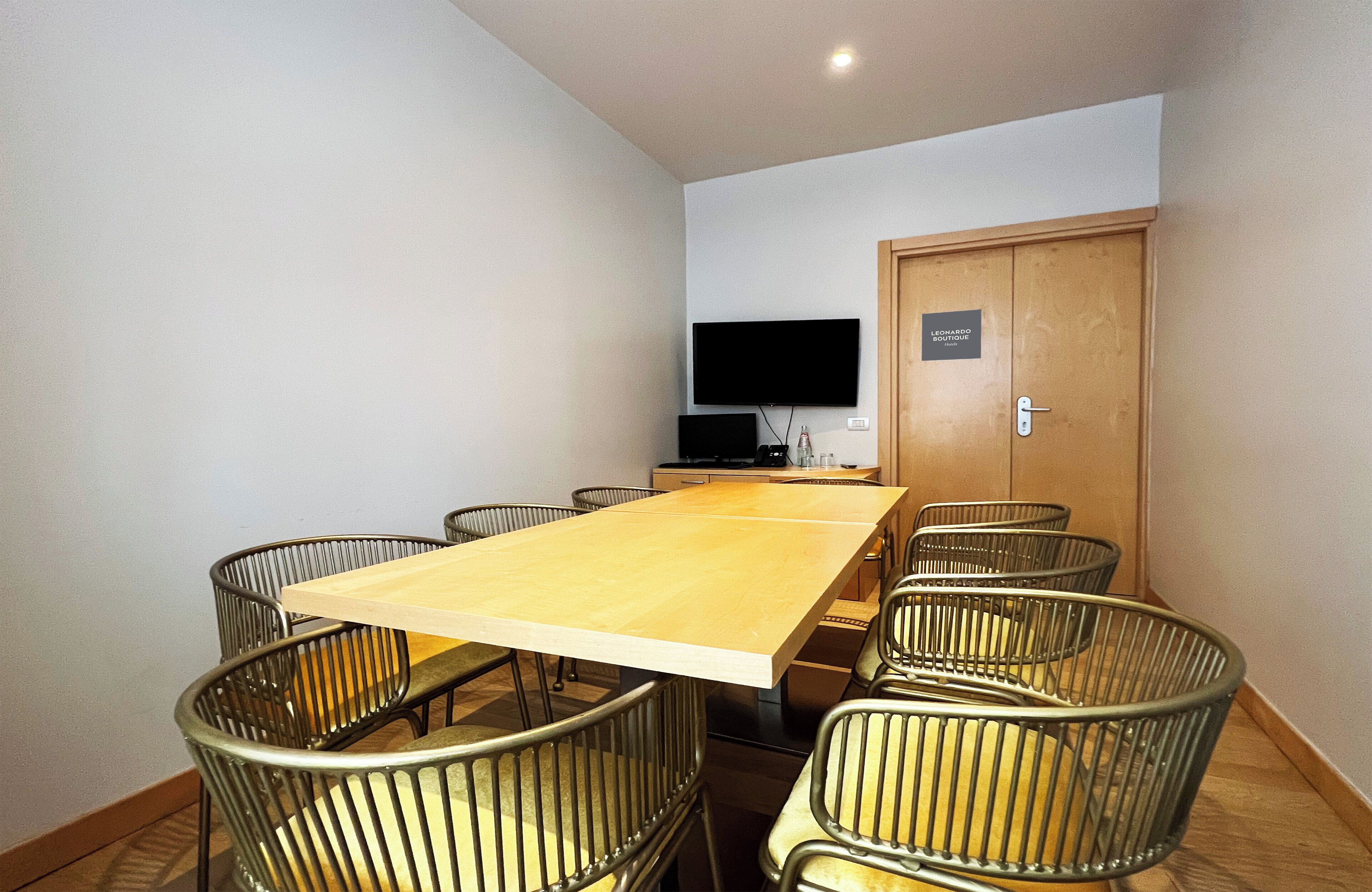 A conference room with a long table, six chairs, a flat-screen TV, and a door labeled 'LEONARDO BOUTIQUE Hotel'.
