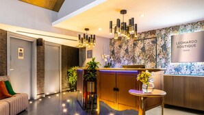 A modern hotel lobby with a reception desk, a sofa, a small table with a vase of flowers, and a wall-mounted sign for the hotel. - Leonardo Boutique Hotel Rome Termini (Rome)