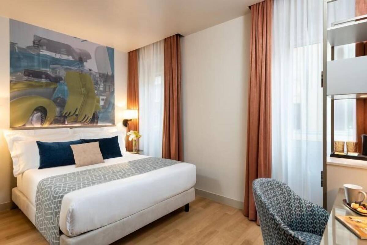 Una habitació d'hotel amb un llit, una cadira, una taula amb una tassa i un quadre a la paret.
