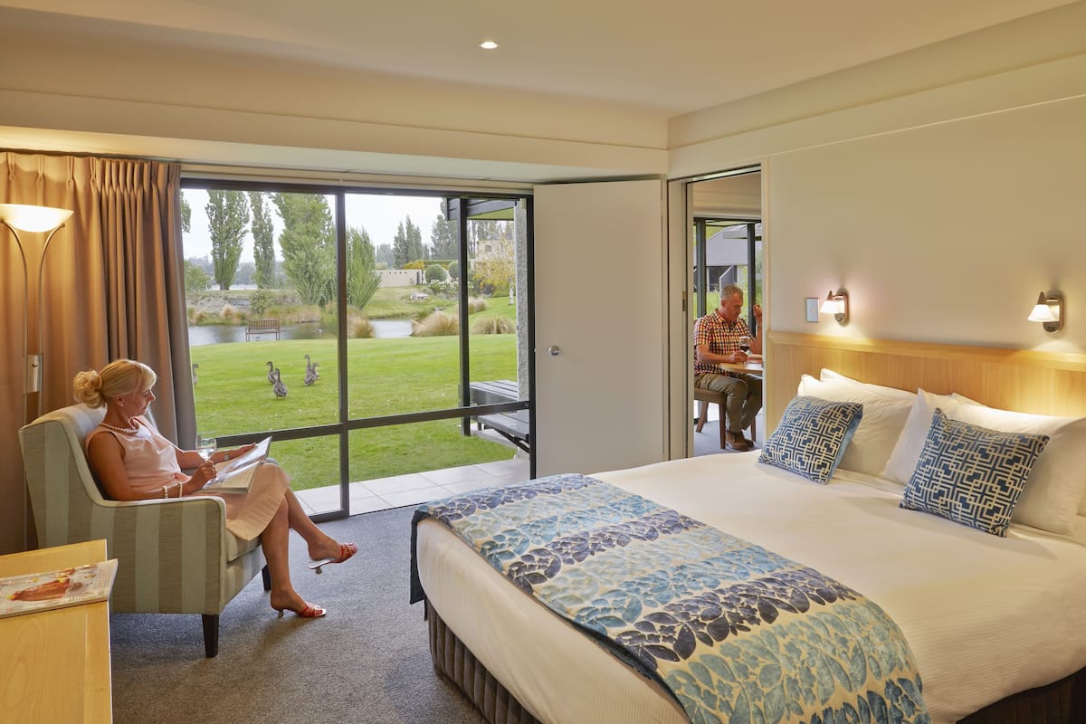 Ein Hotelzimmer mit Bett, Sessel, Tisch und Blick auf Garten und See.