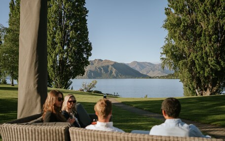 A group of people seated on wicker furniture facing a lake with mountains in the background.