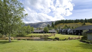 A golf course with a pond, surrounded by buildings with dark roofs and a mountainous backdrop. - Edgewater (Wanaka)