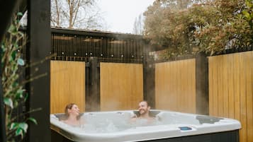 Un bain à remous où se trouvent deux personnes, entouré de clôtures en bois et de plantes.