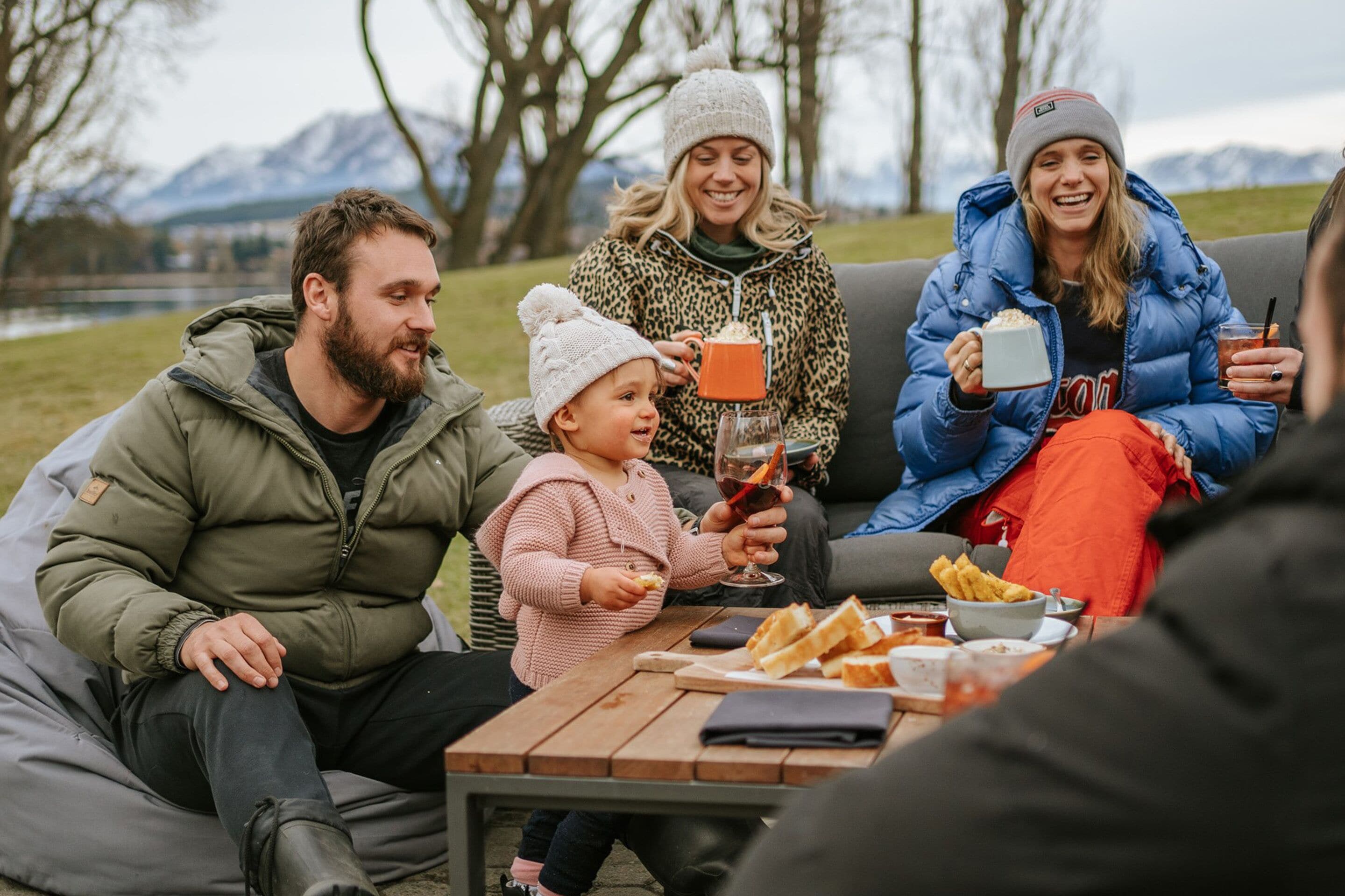 Una familia de cuatro personas sentada en un sofá al aire libre, con una mesa frente a ellas, disfrutando de una comida y bebidas.