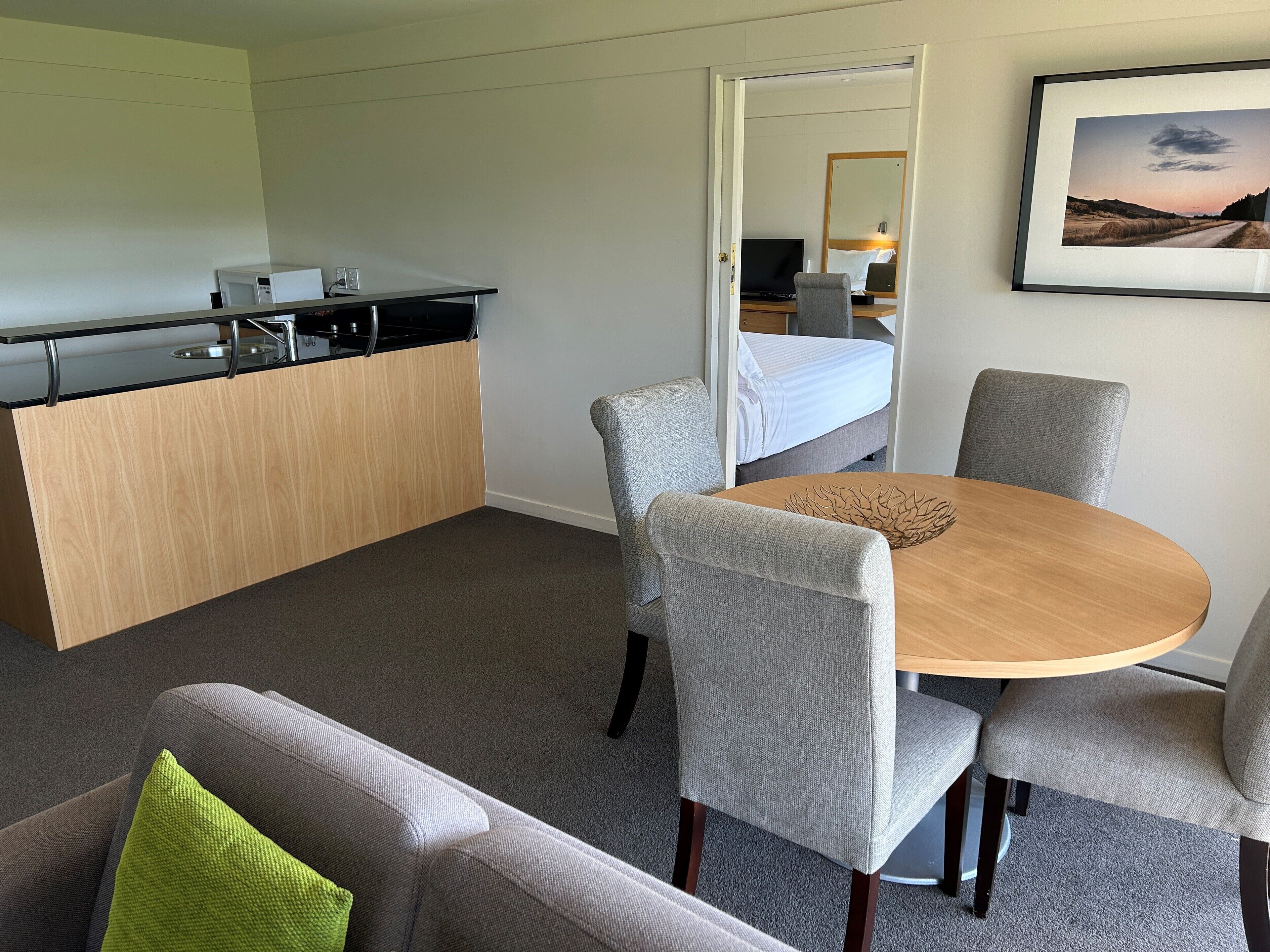 A compact hotel room with a kitchenette, a dining area with a round table and four chairs, a sofa with a green pillow, and a bed with a white headboard.