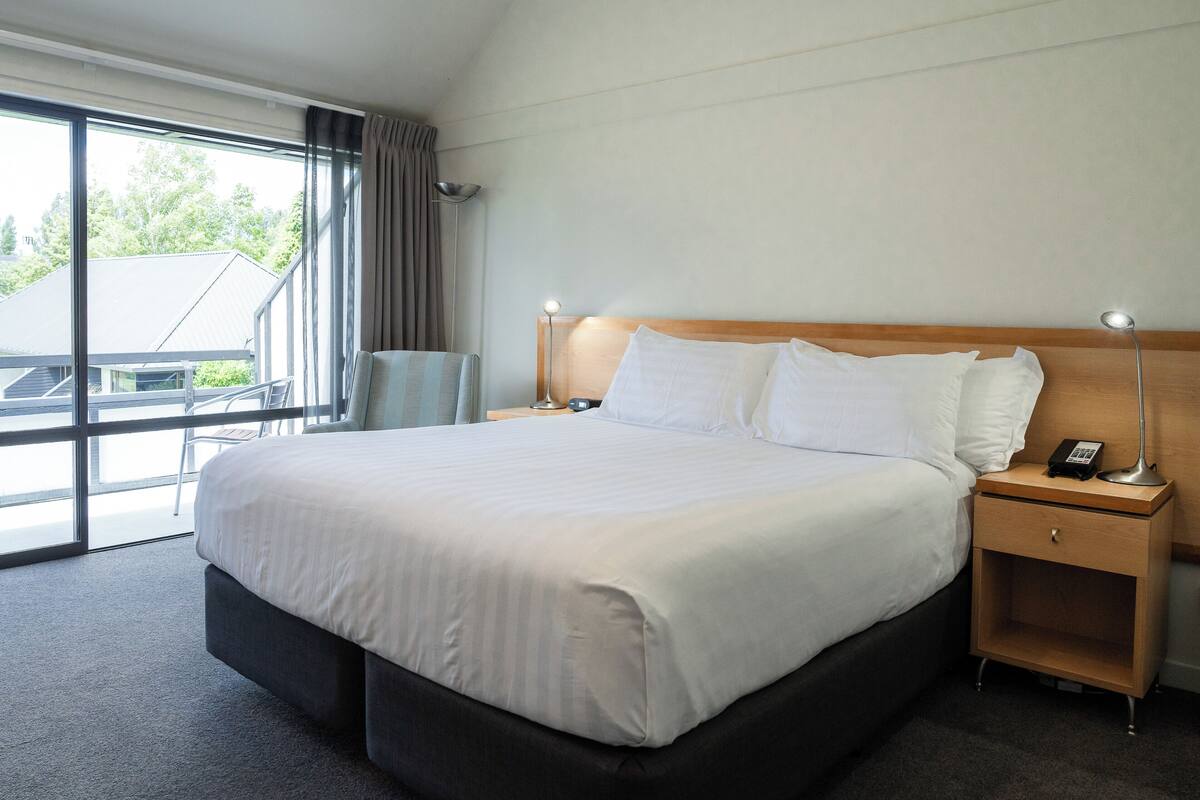 Ein Hotelzimmer mit einem großen Bett, einem Nachttisch mit Lampe und Blick durch ein großes Fenster.