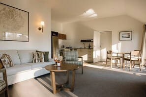 A modern living room with a sofa, a coffee table, and a dining area with chairs. There is a kitchen in the background with cabinets and a sink.