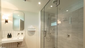 A modern bathroom with a glass-enclosed shower, a white sink with a mirror above, and a small shelf with toiletries.