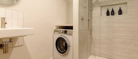 A compact bathroom with a washing machine, a sink, and a glass-enclosed shower.