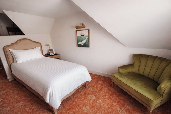 A bedroom with a bed, a nightstand, a green armchair, a painting, and a sloped ceiling.