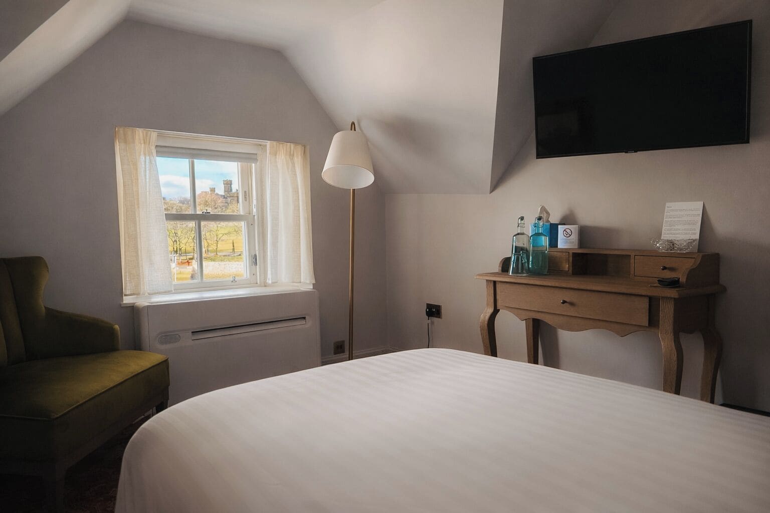 Ein Schlafzimmer mit Bett, Schreibtisch, Stuhl, einem Fenster mit Vorhängen, einer Stehlampe und einem an der Wand montierten Flachbildfernseher.