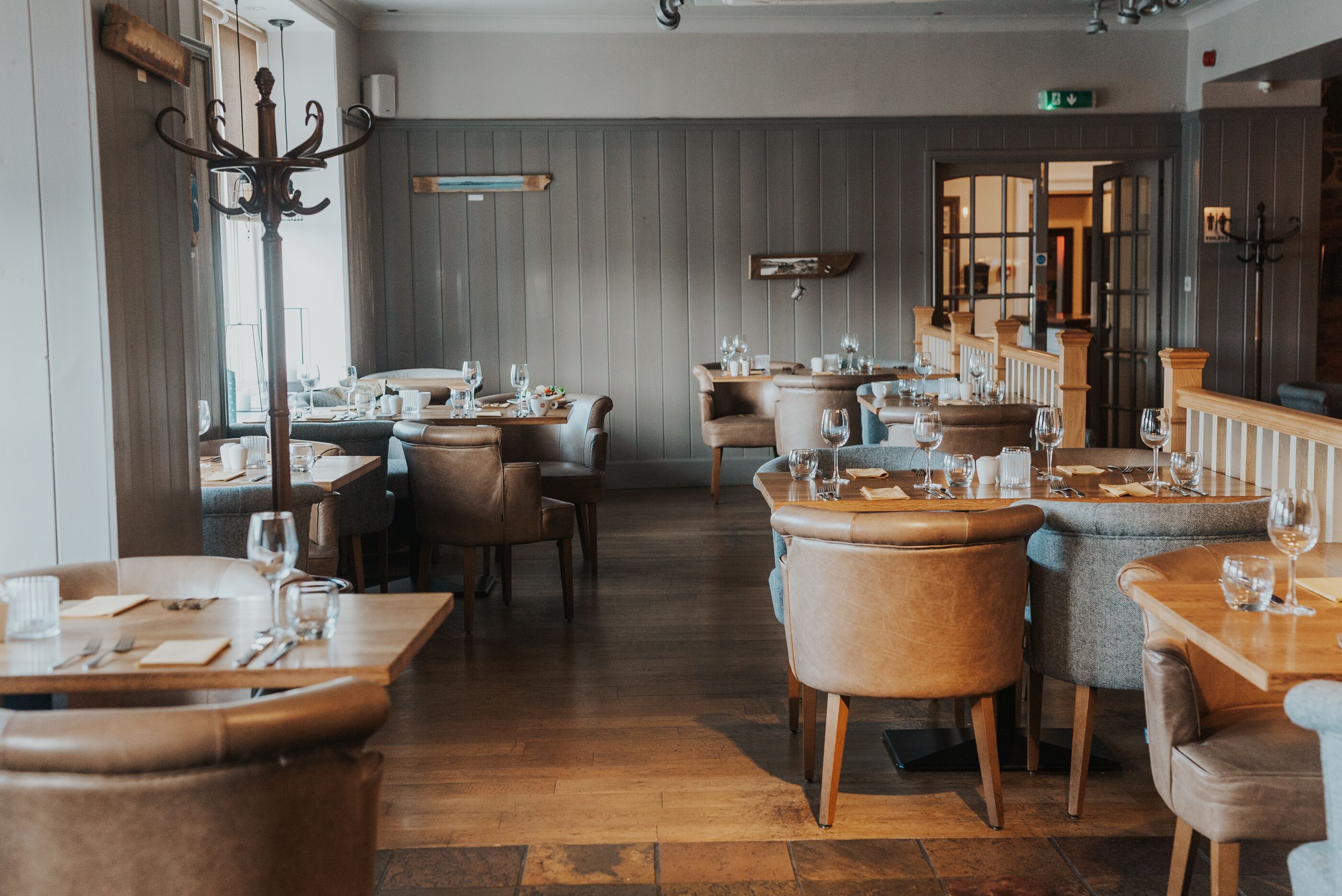 Un restaurant avec des tables et des chaises en bois, un plafond haut et une vue sur l’extérieur grâce à une porte ouverte.