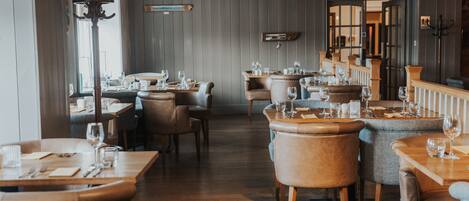 Un restaurant avec des tables et des chaises en bois, un plafond haut et une vue sur l’extérieur grâce à une porte ouverte.