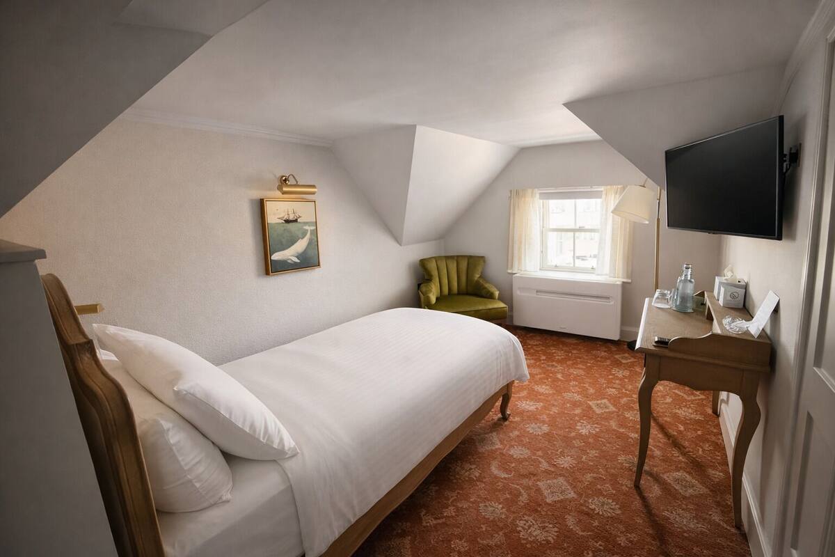 A single bed with white linens, a green armchair, a desk with a lamp, a flat-screen TV, and a window with curtains.