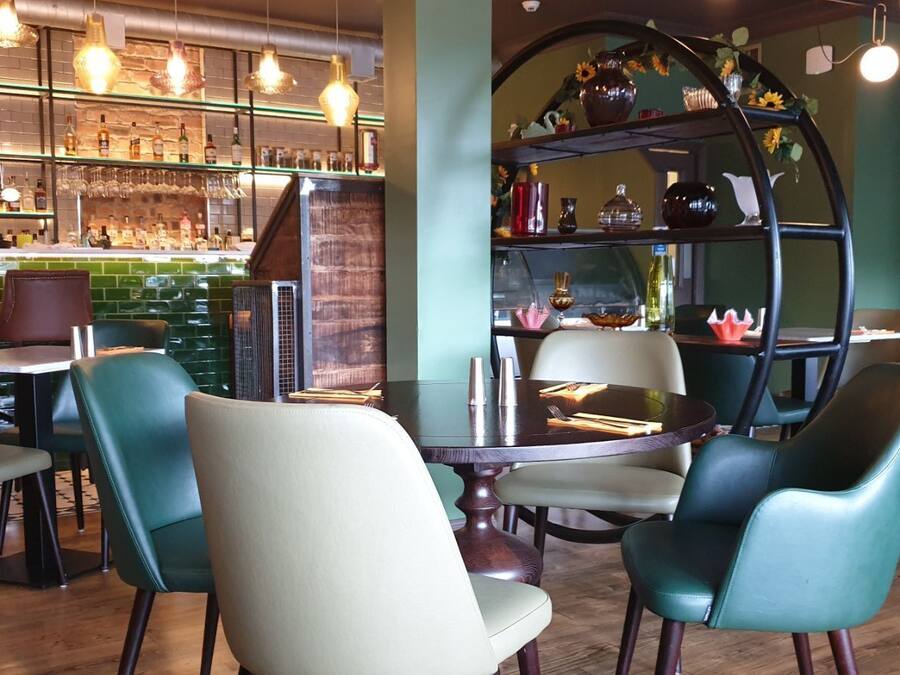 A bar area with a brick wall, shelves with bottles, and a counter with a cash register. There are chairs and tables in the dining area.