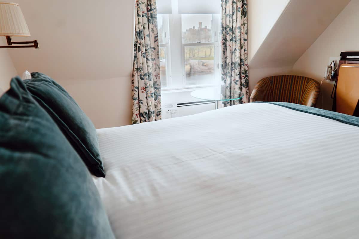 A neatly made bed with a pillow, a chair, and a lamp in a room with a window and curtains.