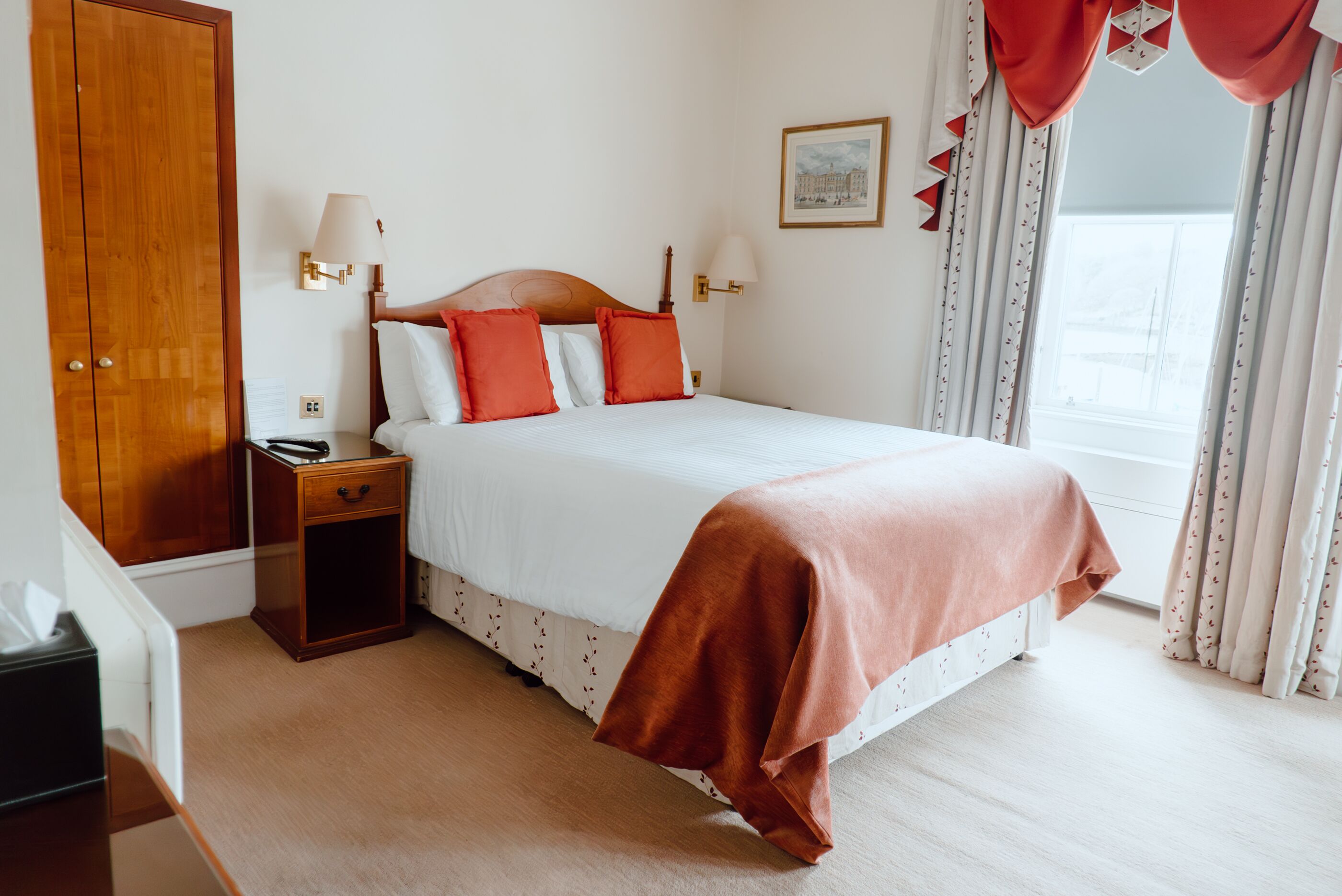 A hotel room with a wooden wardrobe, a bedside table with a lamp, a bed with white bedding and red pillows, and a window with curtains.