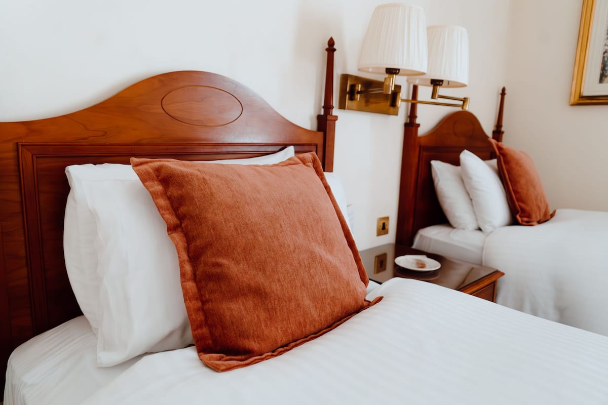 A hotel room with a wooden headboard, a bed with white bedding and a brown pillow, a bedside table with a lamp, and a small table with a plate and a cup.