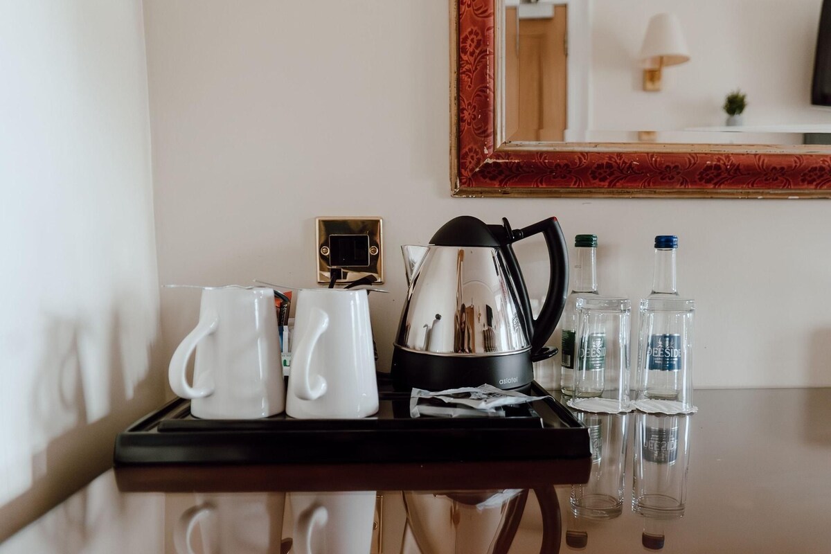 A hotel room's minibar with a coffee maker, two white mugs, a tray, and two glass bottles.