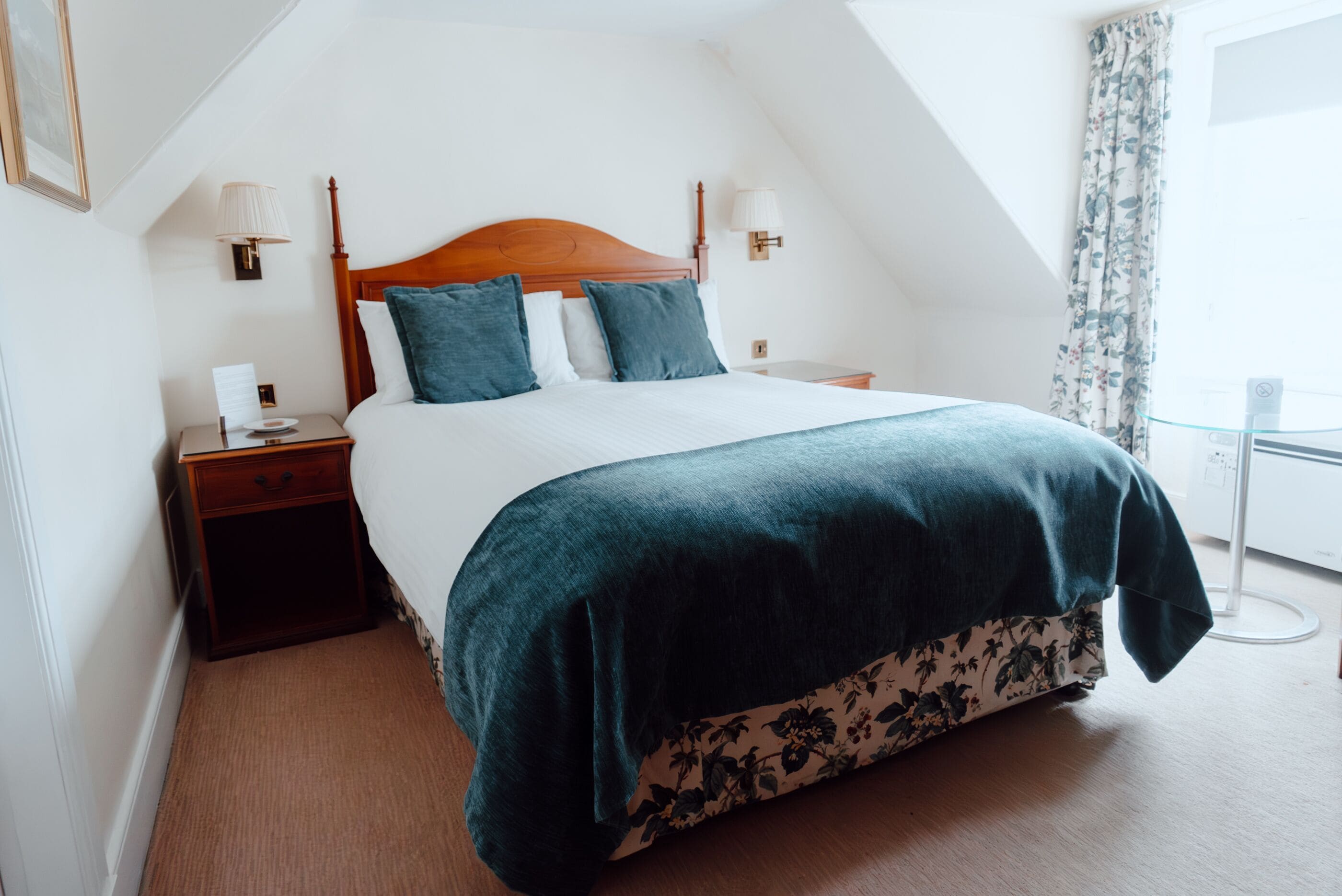 Una cama bien hecha con cabecera de madera, dos mesitas de noche con lámparas y falda de cama estampada con motivos florales.