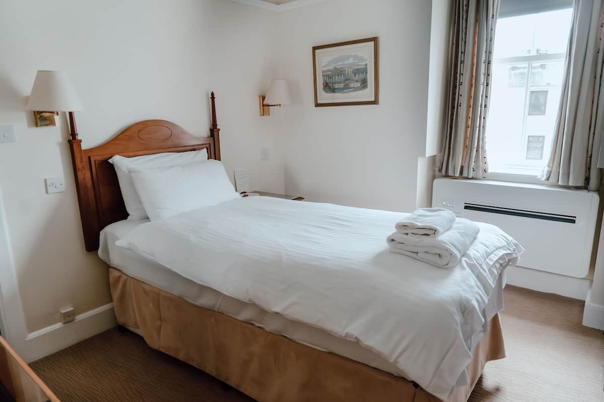 A neatly made bed with white linens and a folded towel on top, a bedside table with a lamp, and a framed picture on the wall.