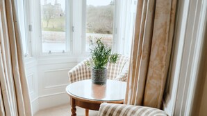 A cozy room with a checkered armchair, a round table with a potted plant, and a view of a building through large windows. - The Royal Hotel (Stornoway)
