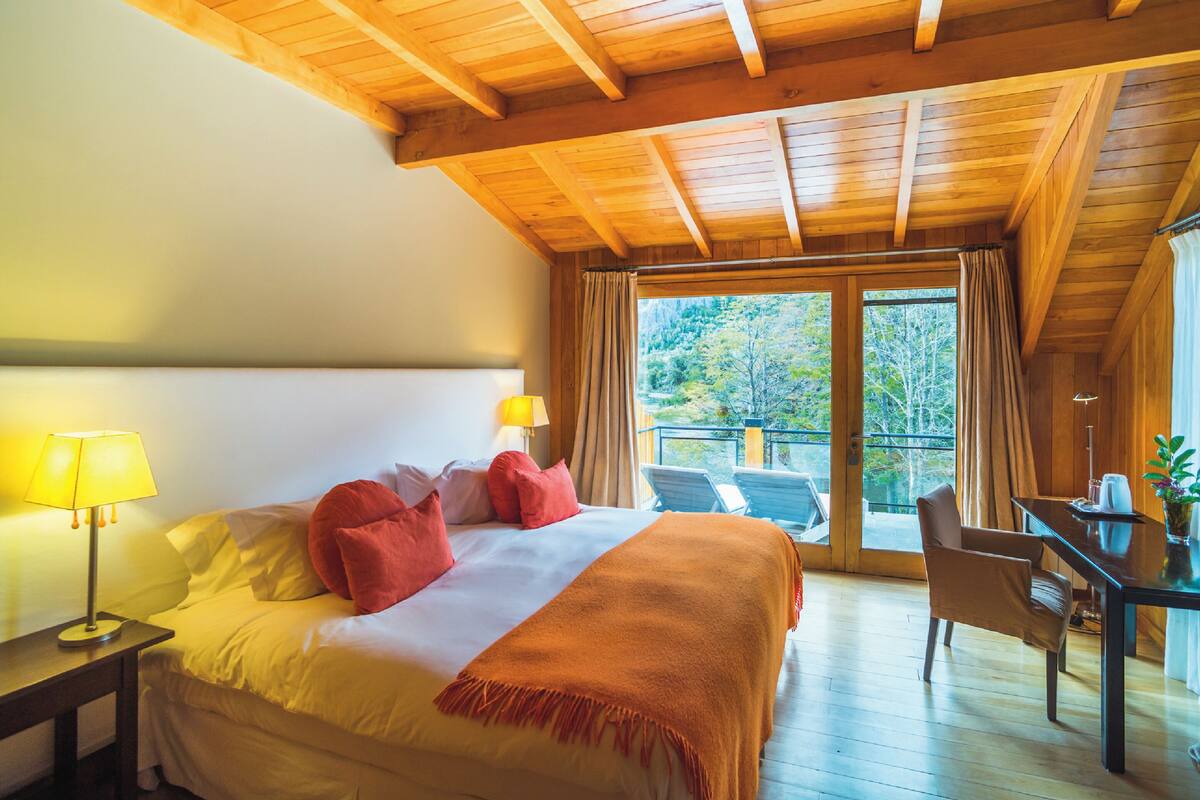 A bedroom with a bed, two bedside lamps, a desk, and a chair. There is a balcony with a view of trees.