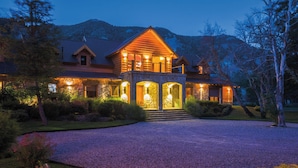 A large, two-story log cabin with a stone entrance and multiple windows, surrounded by a gravel driveway and landscaped garden. - Rio Hermoso Hotel De Montaña (San Martín de los Andes)