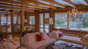 A cozy room with a wooden ceiling, white sofas, a coffee table with books, and a view of a river outside. - Rio Hermoso Hotel De Montaña (San Martín de los Andes)