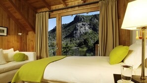 A bedroom with a bed, a bedside table with a lamp, a glass of water, and a view of a mountain and waterfall through a window. - Rio Hermoso Hotel De Montaña (San Martín de los Andes)