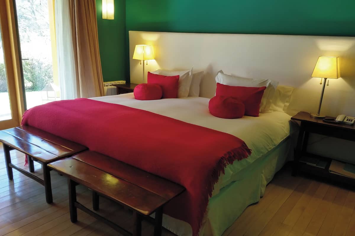 A hotel room with a large bed, two bedside lamps, a bench, and a view of the outdoors.