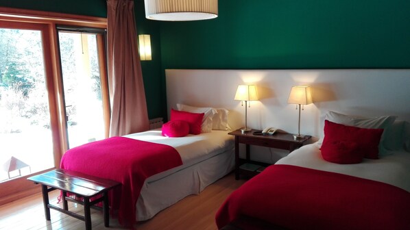 Une chambre d’hôtel avec deux lits, chacun agrémenté d’oreillers rouges et de draps blancs, un banc en bois, une table de chevet avec une lampe, et une vue sur la verdure grâce à une grande fenêtre.