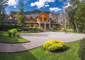 A large, multi-story wooden house with a stone foundation, surrounded by well-maintained gardens and a gravel driveway.