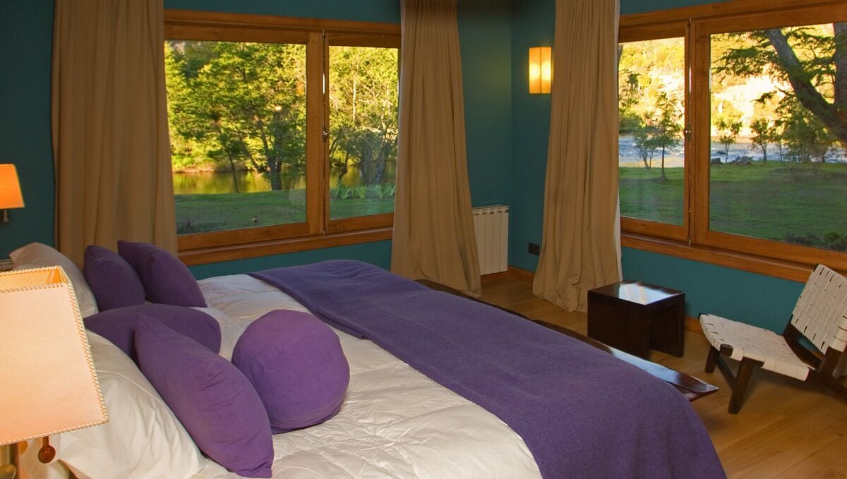 Quarto com cama grande, duas mesas de cabeceira, uma cadeira e vista para um lago através de janelas amplas.