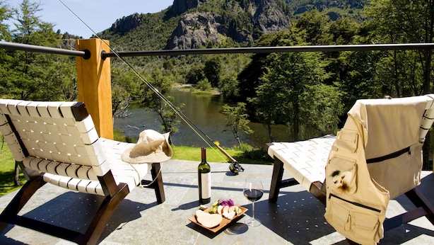 A terrace with two chairs, a table, a bottle of wine, a plate with food, and a fishing rod overlooking a river and mountains. - Rio Hermoso Hotel De Montaña (San Martín de los Andes)