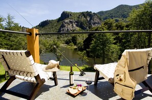 Une terrasse avec deux chaises, une table, une bouteille de vin, une assiette de nourriture et une canne à pêche, le tout donnant sur une rivière et des montagnes.
