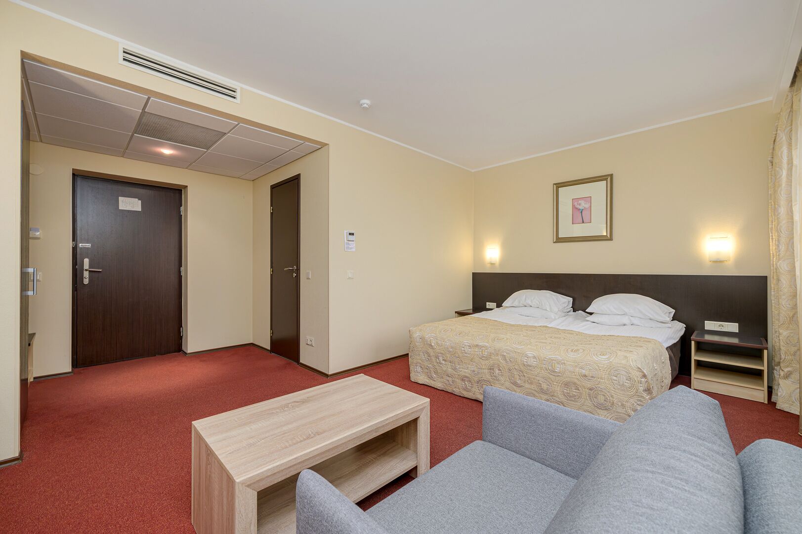 Ett hotellrum med en säng, ett nattduksbord, ett litet bord i trä, en grå soffa och en röd matta.