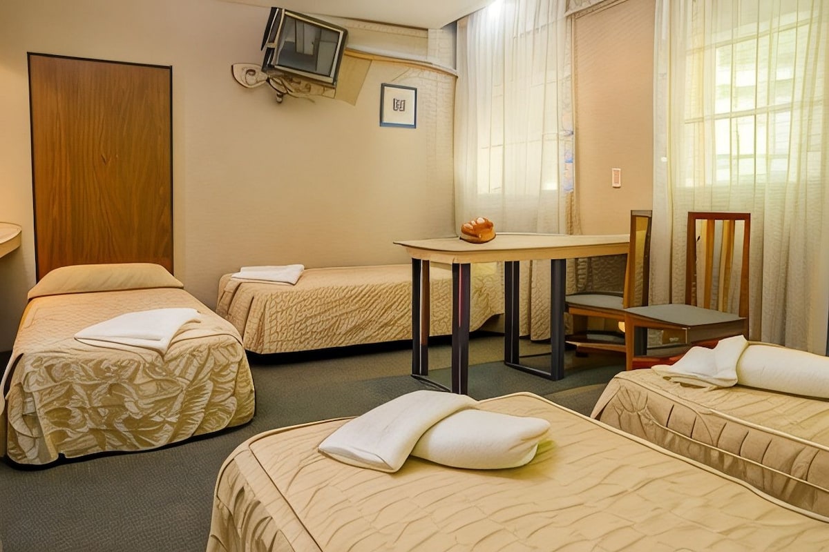 A hotel room with two beds, a small table, a chair, a television, and a window with curtains.