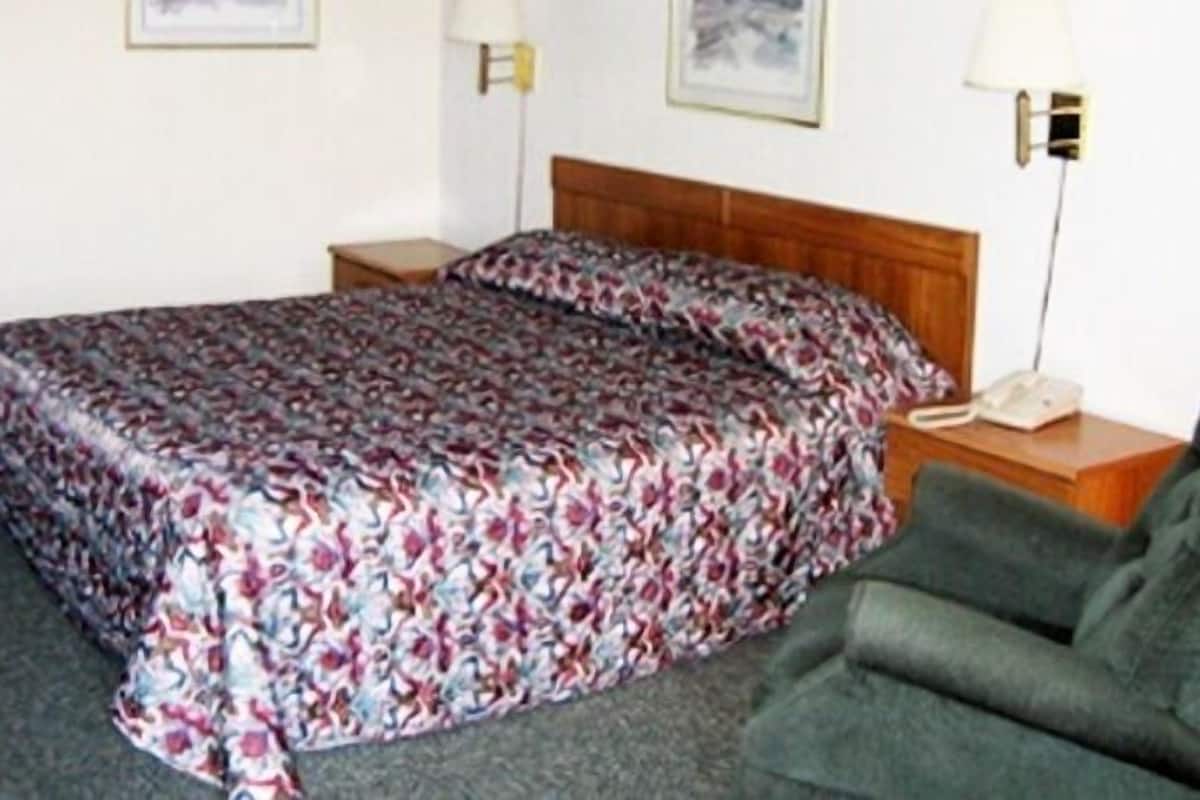 A hotel room with a bed, a chair, a nightstand with a phone, and a lamp.
