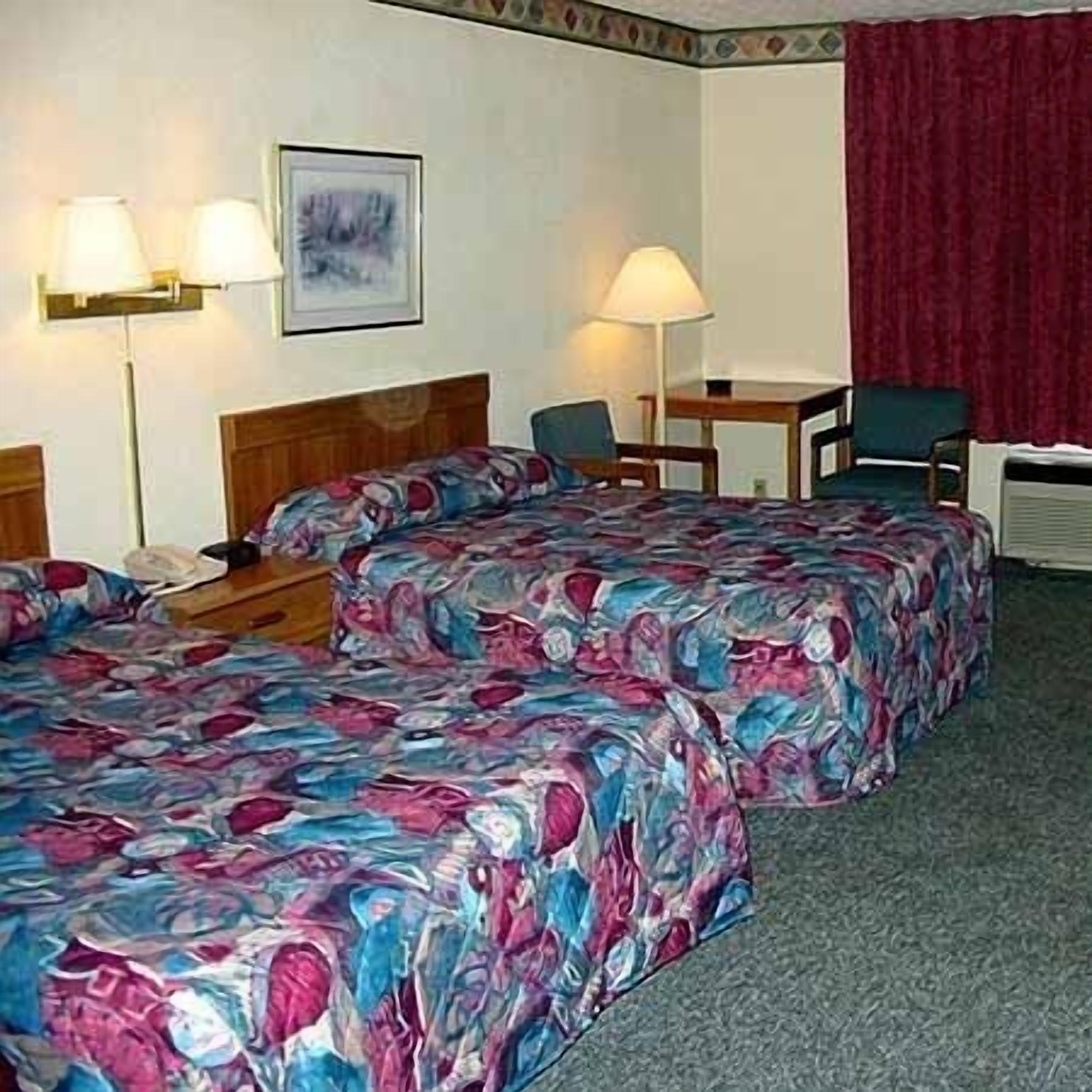 A hotel room with two beds, a desk, two chairs, a lamp, and a window with a red curtain.