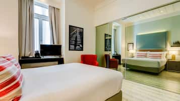 Quarto de hotel com uma cama grande, uma escrivaninha com televisão, uma cadeira vermelha, outra cama e um espelho refletindo o quarto.