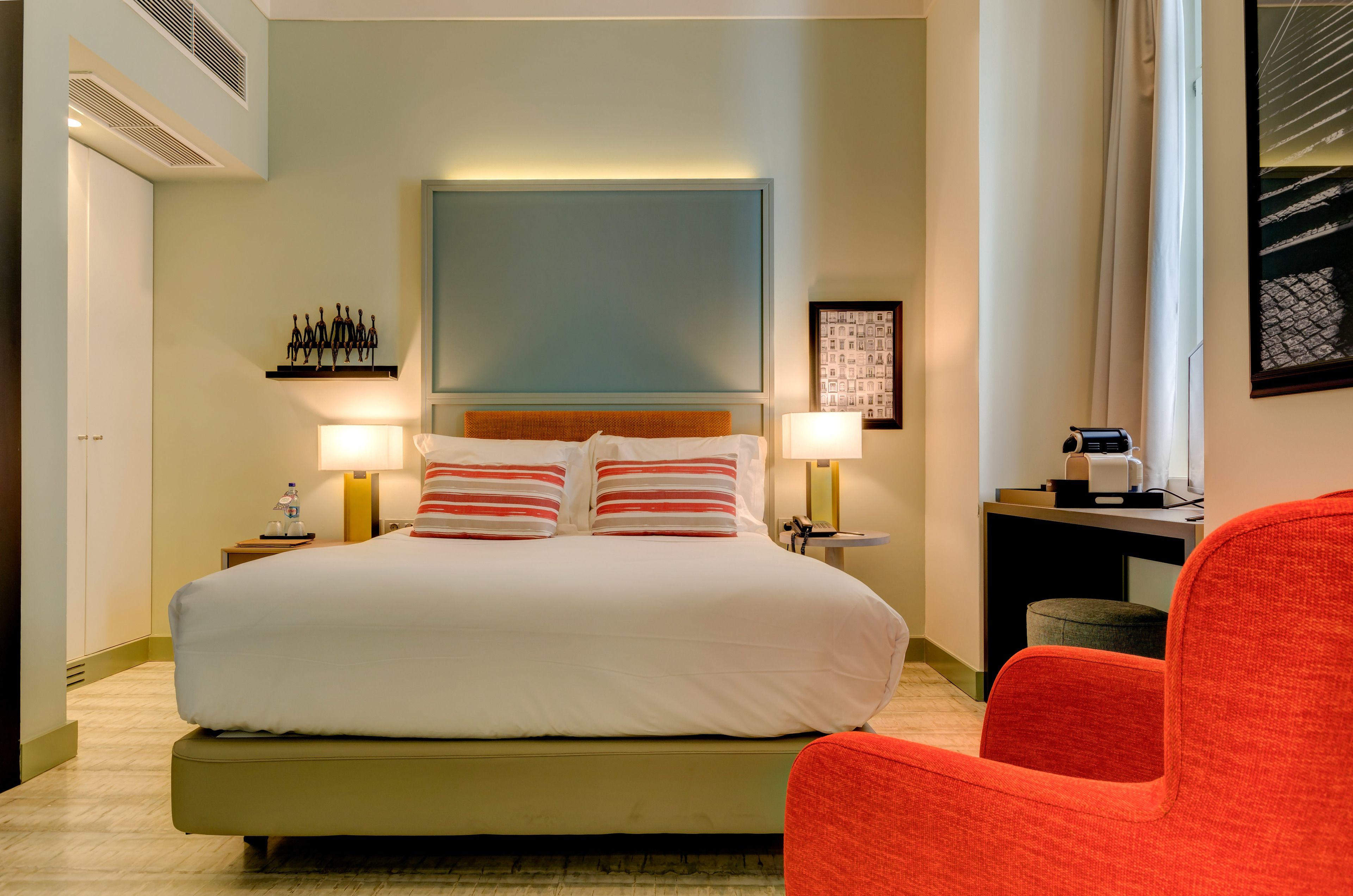 A hotel room with a large bed, two bedside lamps, a nightstand with a water bottle, a desk with a lamp, and a red chair.