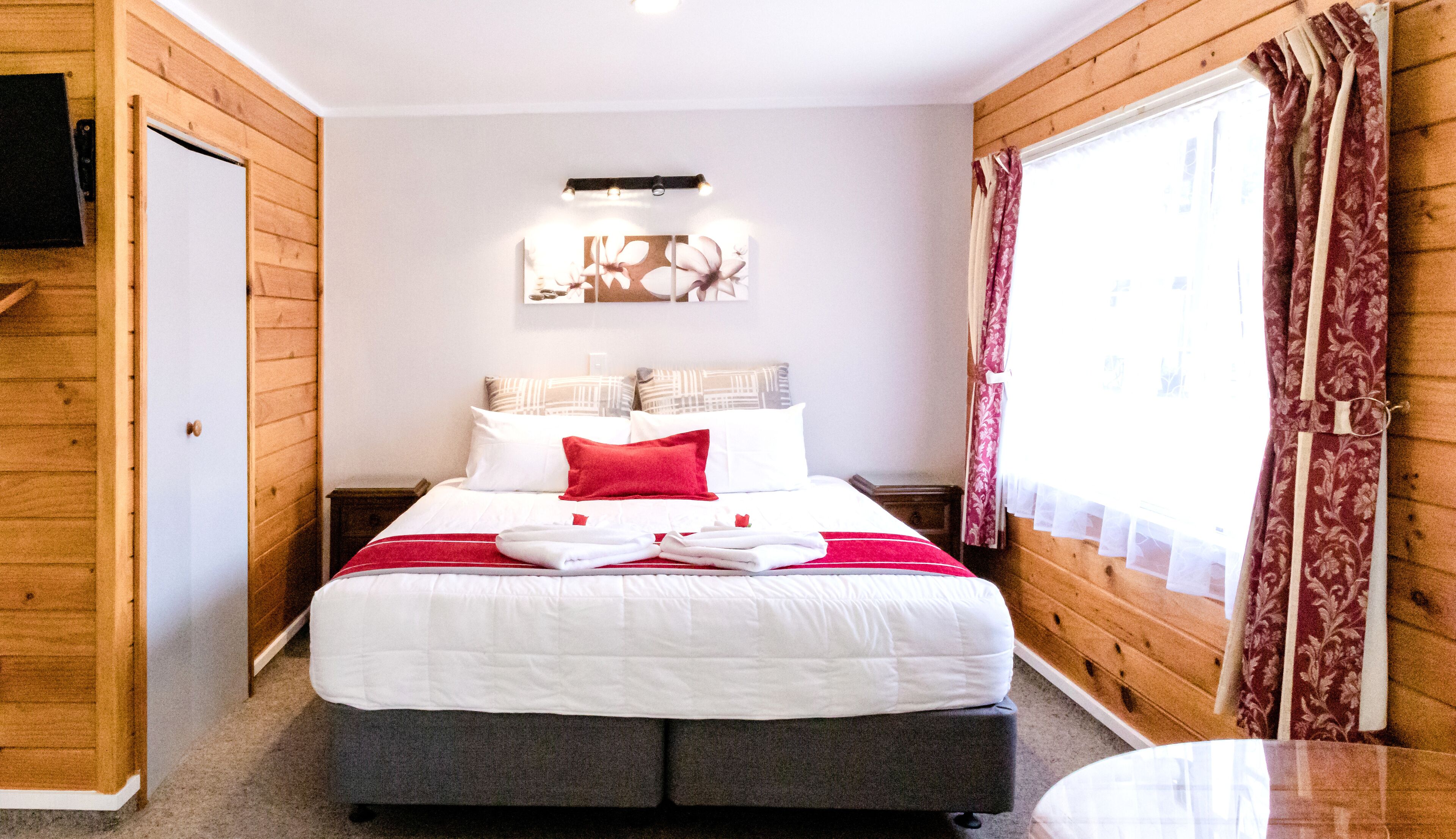 A bedroom with a bed, a nightstand, a wall-mounted TV, and a window with curtains.